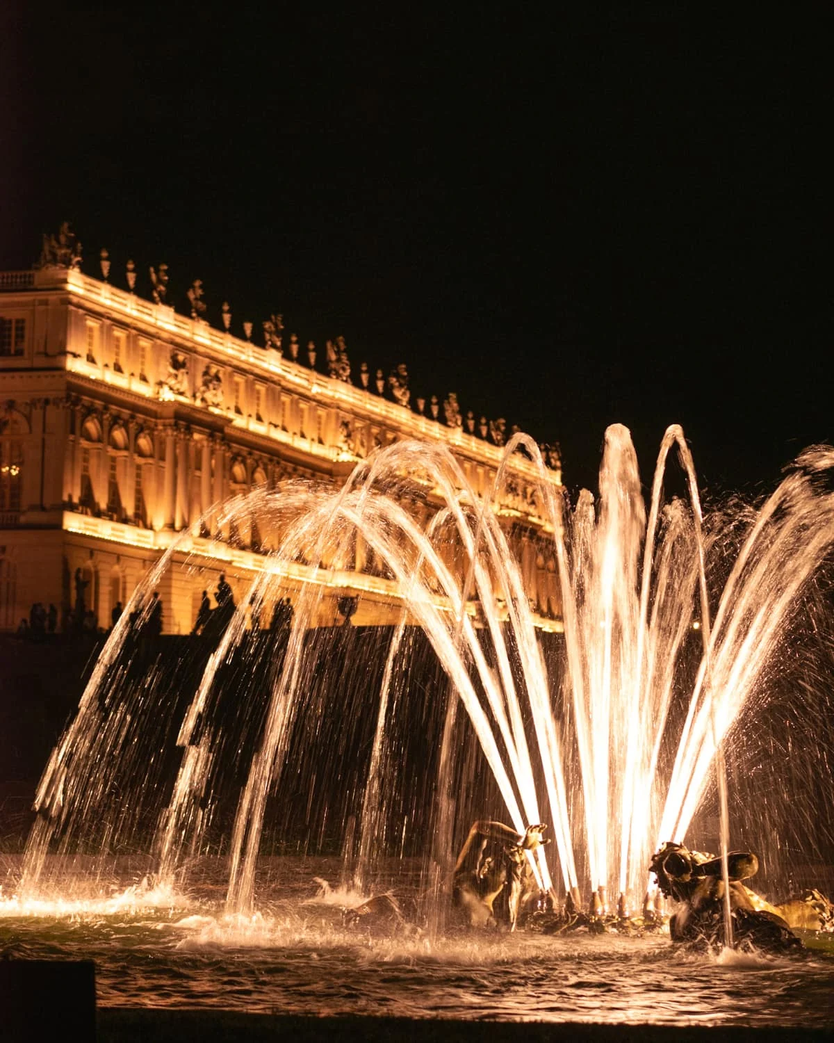 A fountain with the backdrop of chateau de Versailles at night