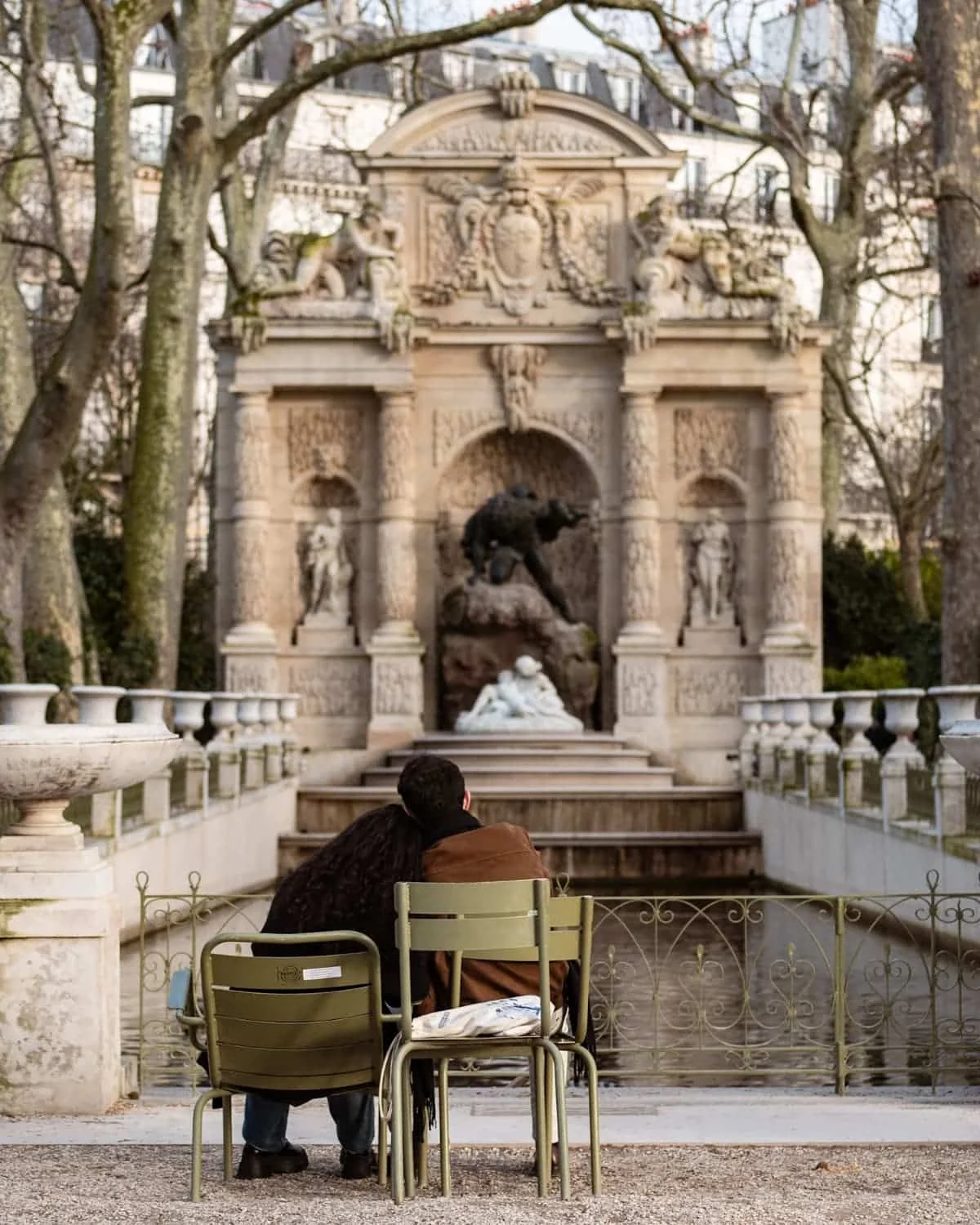 A couple sitting on chairs in front of the La fontaine Médicis